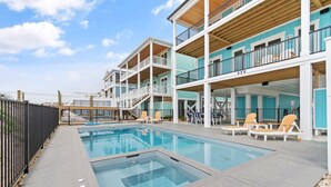 A pool area with a wooden deck, lounge chairs, and a multi-level building with balconies.