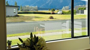 A room with a large window offering a view of a road, a small building, and mountains in the distance. - Brand New 3-bedroom house with lake and mountain views, AC, lots of TVs, Te Anau (Te Anau)
