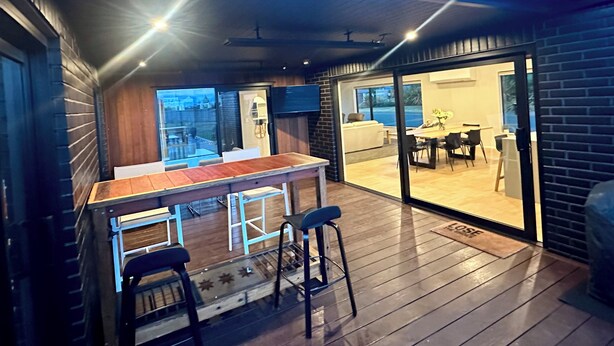 A modern outdoor patio with a wooden table and bar stools, a glass door leading to a dining area, and a view of the surrounding landscape. - Brand New 3-bedroom house with lake and mountain views, AC, lots of TVs, Te Anau (Te Anau)