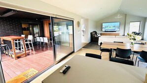 A modern living room with a large glass door leading to a dining area with a wooden table and chairs. There is a television mounted on the wall, a sofa, and a decorative vase with flowers. - Brand New 3-bedroom house with lake and mountain views, AC, lots of TVs, Te Anau (Te Anau)