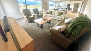 A spacious living room with a large window, a wooden coffee table, and a sofa with a green throw. - Brand New 3-bedroom house with lake and mountain views, AC, lots of TVs, Te Anau (Te Anau)