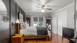 Un dormitorio con una cama de madera, ventilador de techo, mesita de noche, cómoda y un ventanal grande.