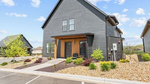 A modern two-story building with a dark exterior, a front porch, and a landscaped front yard with various plants and shrubs.