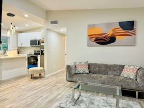A modern living room with a gray sofa, ottoman, and glass coffee table. A kitchen with white cabinets and a microwave is visible in the background.