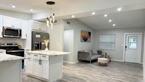 Una cocina moderna con armarios blancos, una isla central y un rincón comedor con un sofá y reposapiés grises.