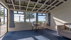 A room with a large window, a table with chairs, a sofa, and a carpeted floor. - Waterfront | Great Views | Affordable | Unit 3 - KK61643 (Kaikoura)