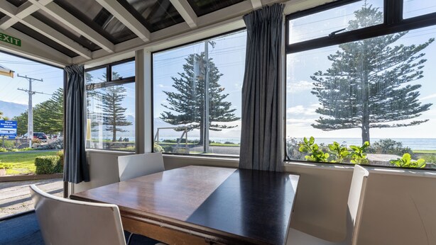A conference room with a large table and white chairs, large windows with curtains, and a view of trees and a body of water. - Waterfront | Great Views | Affordable | Unit 3 - KK61643 (Kaikoura)