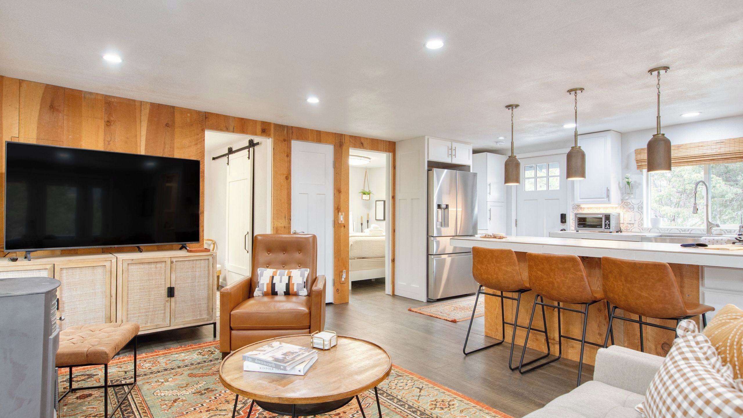 A modern living room with a large TV, leather armchair, coffee table, and a kitchen area with bar stools and a refrigerator.