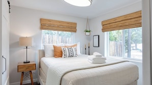 A bedroom with a bed, bedside table, lamp, window with blinds, and a hanging plant.