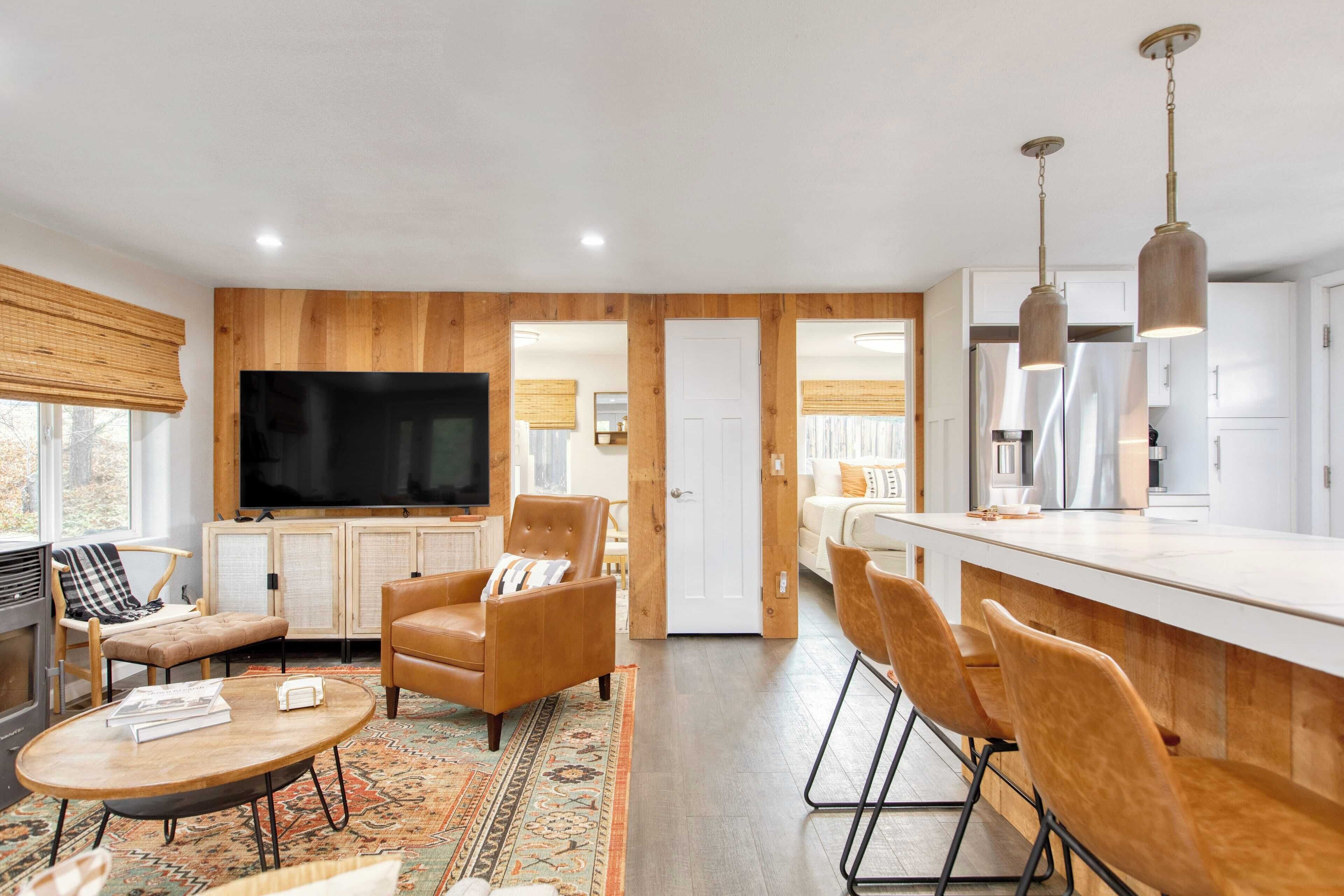 A modern living room with a large TV, leather armchairs, a coffee table, and a kitchen area with bar seating.