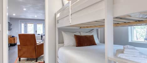 A bunk bed room with white bedding, a brown chair, and a window with daylight.