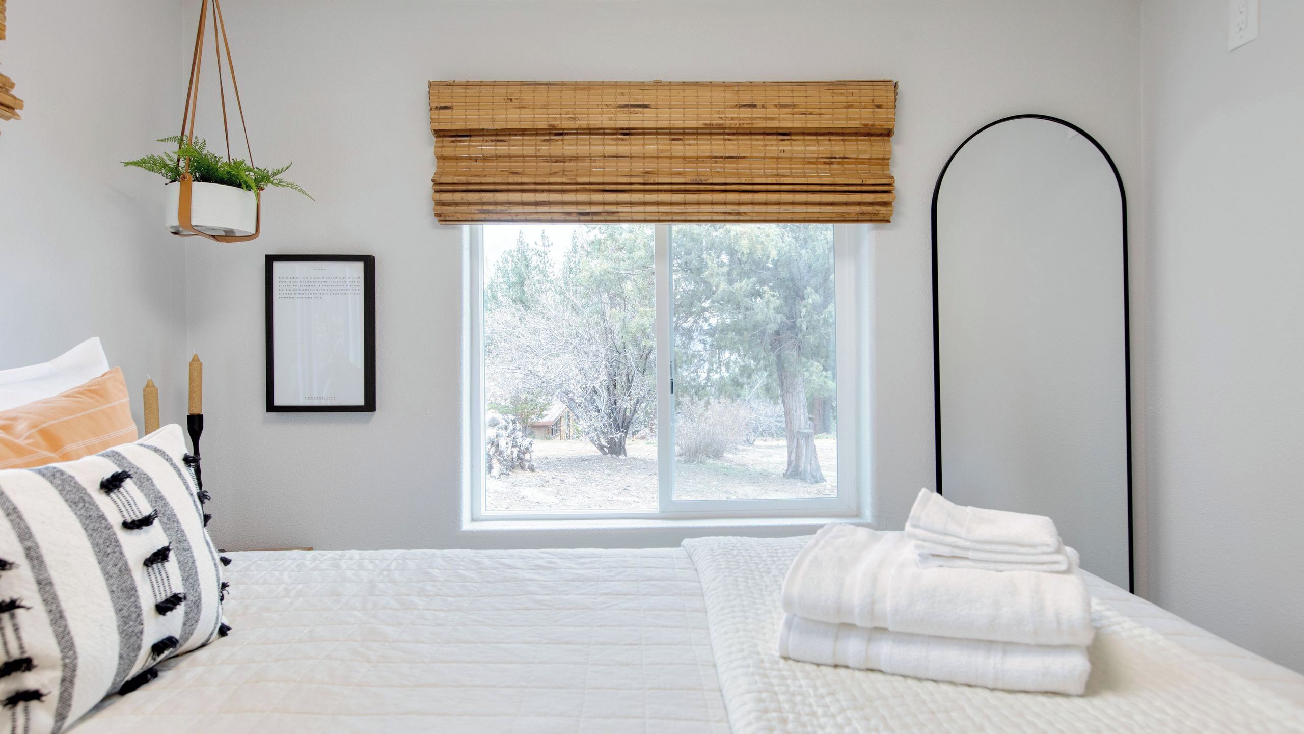 Une chambre à coucher avec un lit, une fenêtre donnant sur des arbres, une plante suspendue et un tableau encadré au mur.