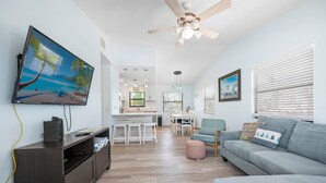 A modern living room with a flat-screen TV, a ceiling fan, a grey sofa, and a dining area in the background.