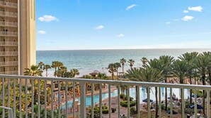 A hotel balcony with a view of the ocean, a pool area with palm trees, and a beach in the background. - Relax at Panama City Beach Balcony, Parking, Walk to Dining (Panama City Beach)