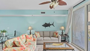 A living room with a ceiling fan, a fish wall decoration, a sofa, a coffee table, and a dining area with chairs and a table. - Relax at Panama City Beach Balcony, Parking, Walk to Dining (Panama City Beach)