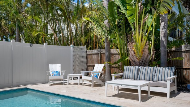 Un espace piscine agrémenté de chaises longues blanches et d’un canapé blanc, le tout entouré d’une clôture en bois et de plantes tropicales.