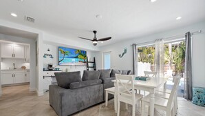 Un salon moderne avec un canapé gris, une table à manger blanche et des chaises. On y trouve un ventilateur de plafond, un tableau représentant une scène de plage et une vue sur l’extérieur grâce à de grandes fenêtres.