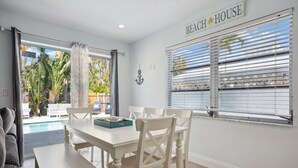 Un coin repas avec une table et des chaises blanches, une enseigne indiquant « MAISON DE PLAGE », et une vue sur une piscine et la végétation environnante.