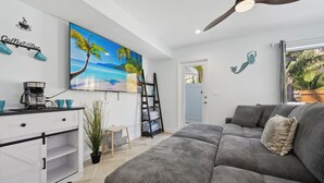 Un salon moderne avec un canapé gris, un coin café, un tableau représentant une plage et un ventilateur de plafond.