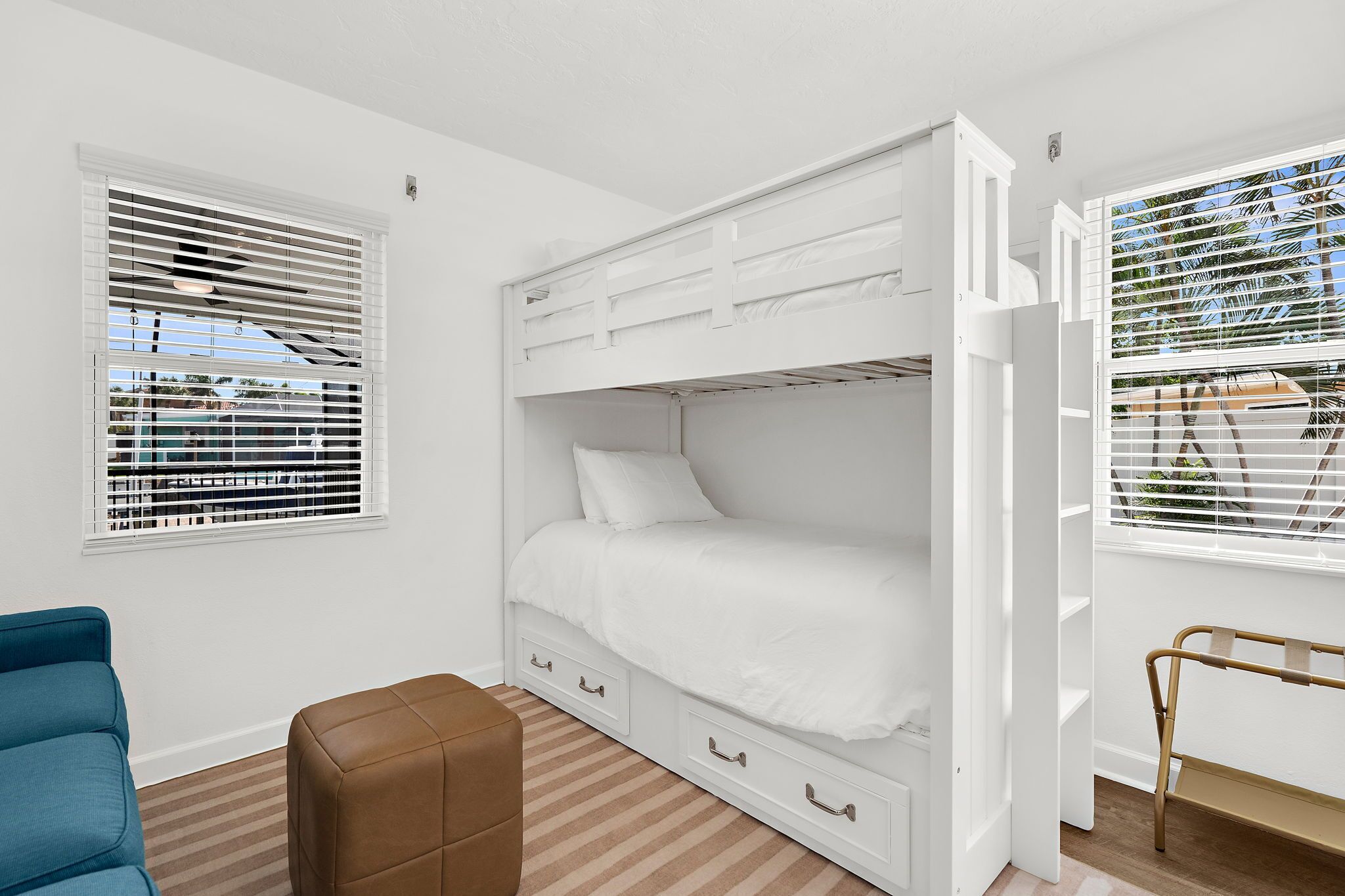 A bunk bed room with white walls, a blue chair, a brown ottoman, and a window with blinds.