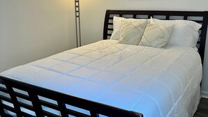 A neatly made bed with a white duvet and decorative pillows, a floor lamp, and a framed abstract painting on the wall.