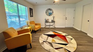 A living room with a round rug, two yellow armchairs, a ceiling fan, a window with blinds, a framed picture, a small table with a globe and candle, and a white door.