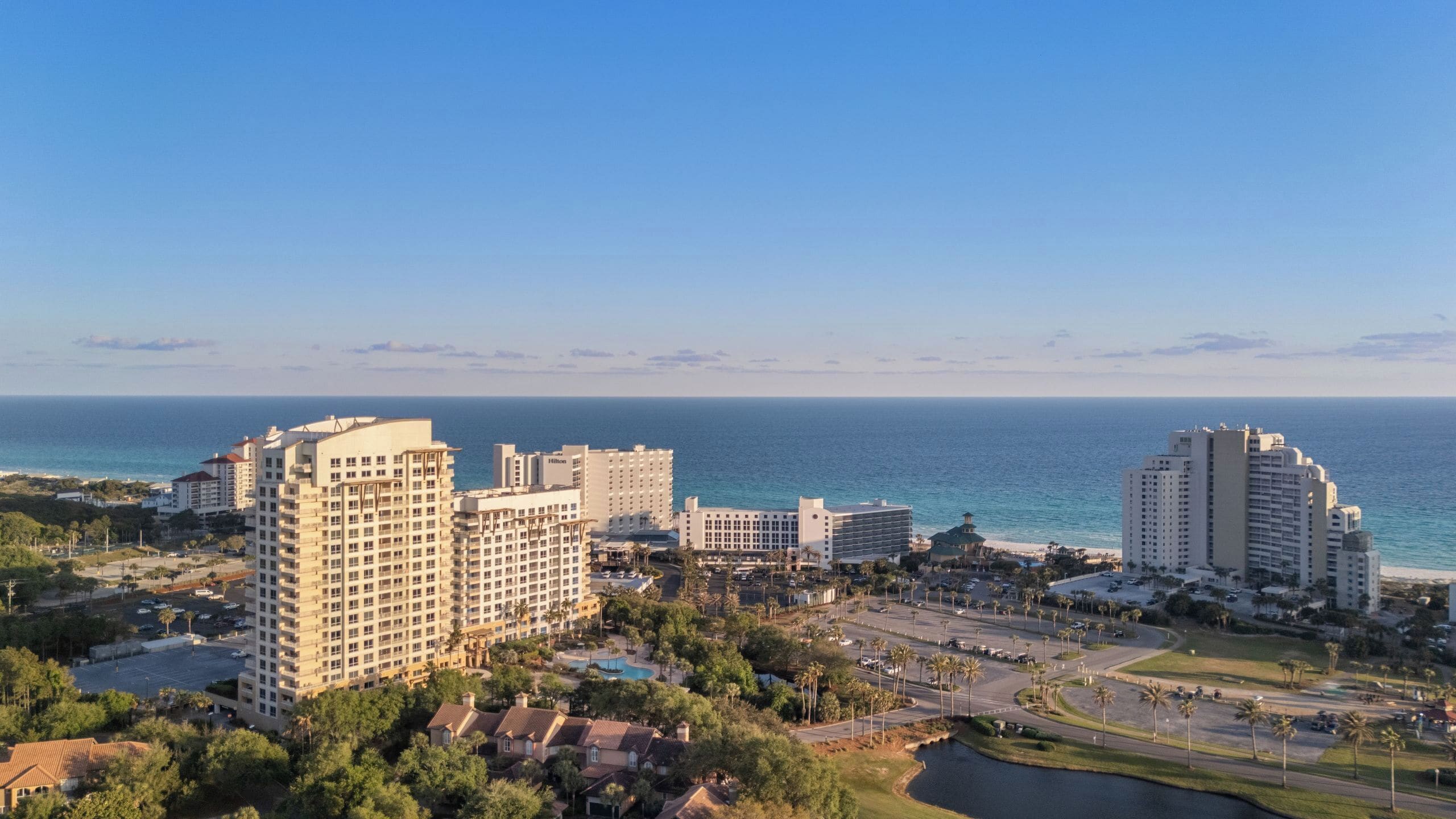 A coastal hotel area with multiple high-rise buildings, a parking lot, and a clear view of the ocean.