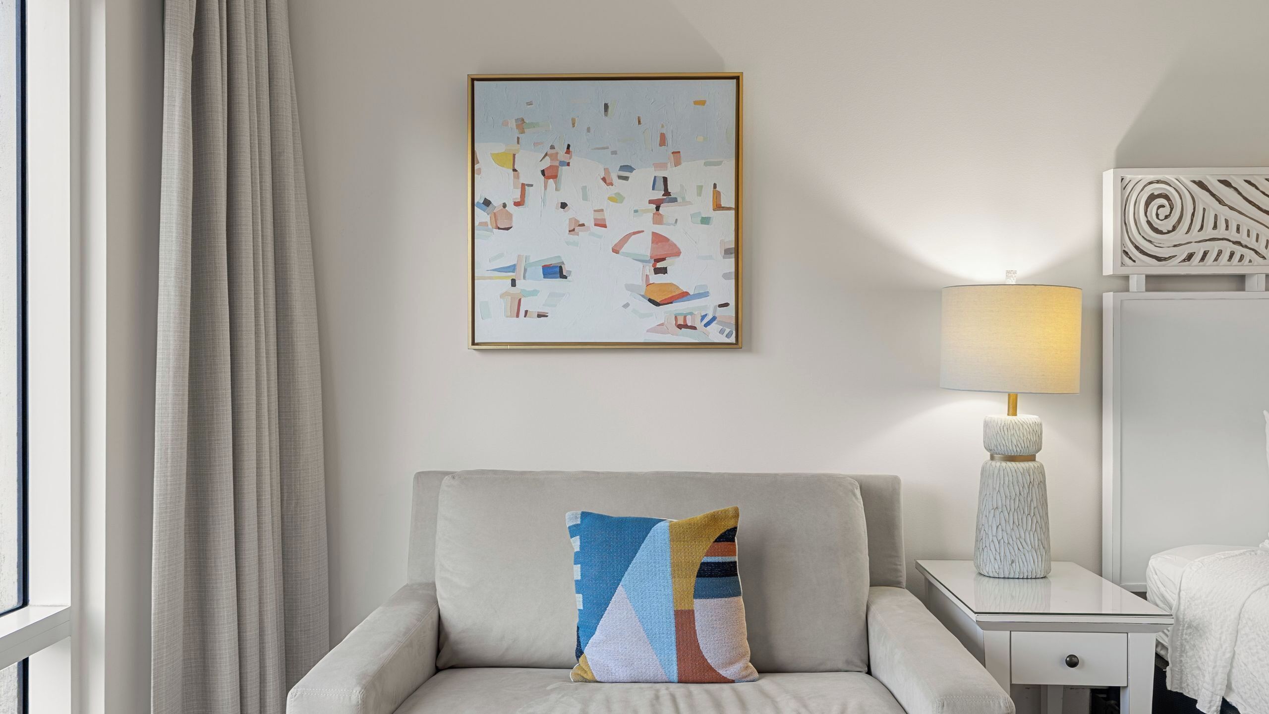 A modern hotel room with a light-colored sofa, a colorful abstract painting, a lamp, and a bed with a white cover.
