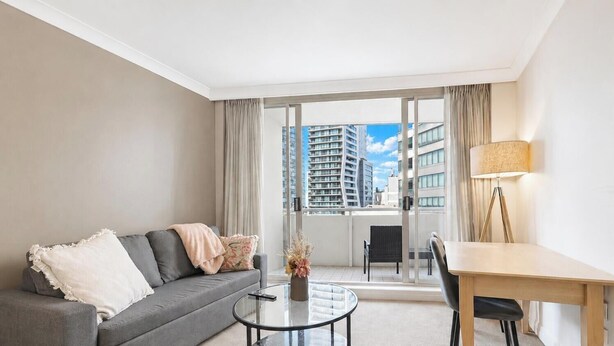 A modern living room with a grey sofa, a glass coffee table, a wooden dining table, and a view of high-rise buildings through large windows. - Cozy Studio with Gym and Pool Near Chatswood Station (Liberty Grove)