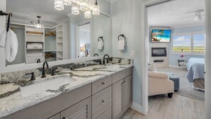 A bathroom with a large mirror, two sinks, and a vanity with drawers. There is a walk-in closet with shelves and a hanging rack. The bathroom has a ceiling fan and a view of a bedroom with a bed and a TV.