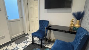 Een kamer met een tapijt met pauwenveren, twee blauwe fauteuils, een zwarte tafel, een gouden decoratieve vaas en een tv aan de muur.
