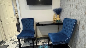 Een kamer met een vloerkleed met pauwenveren, twee blauwe fauteuils, een zwarte televisie aan de muur en een decoratieve vaas.