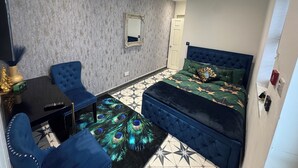 Een slaapkamer met een bed met een gestuikt hoofdbord, een vloerkleed met pauwenprint, een blauwe bank, een kleine tafel en een spiegel aan de muur.