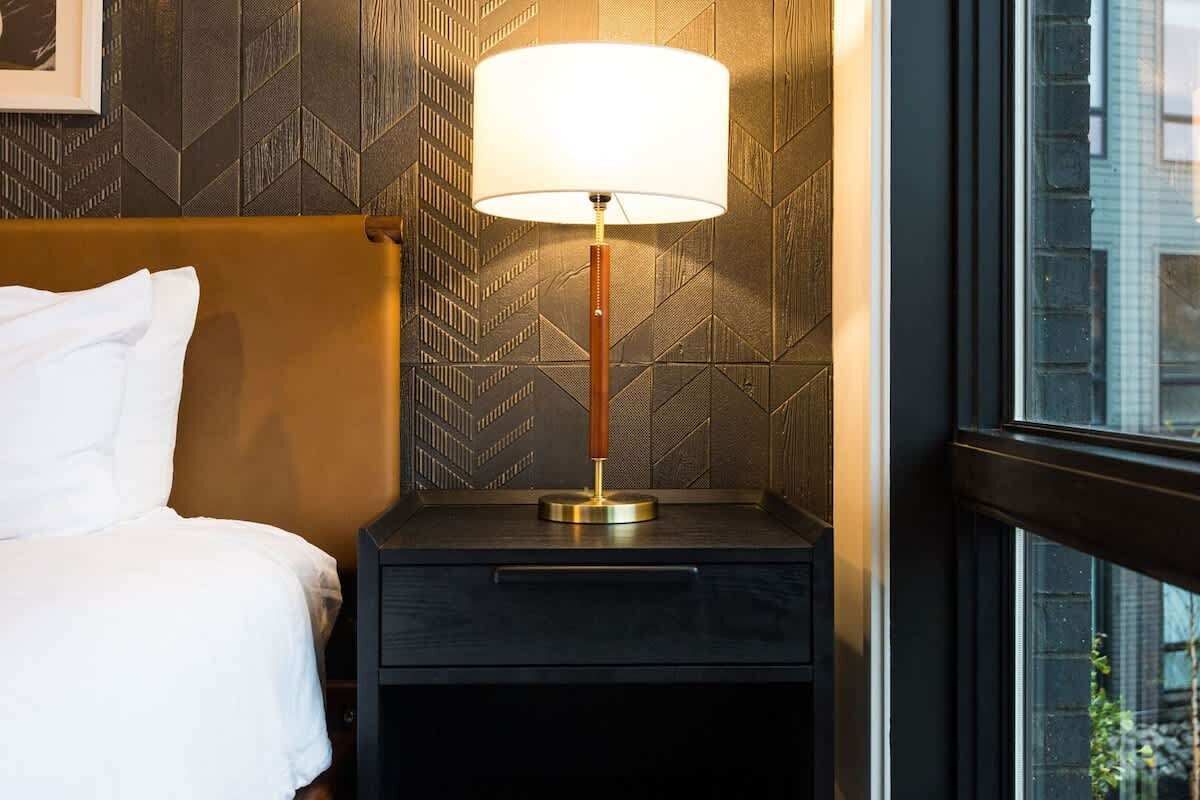 A neatly made bed with white linens, a small bedside table with a lamp, and a patterned wall.