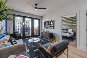 Una sala moderna con un sofá, una mesa de centro y un televisor. Hay un balcón con vistas a edificios.