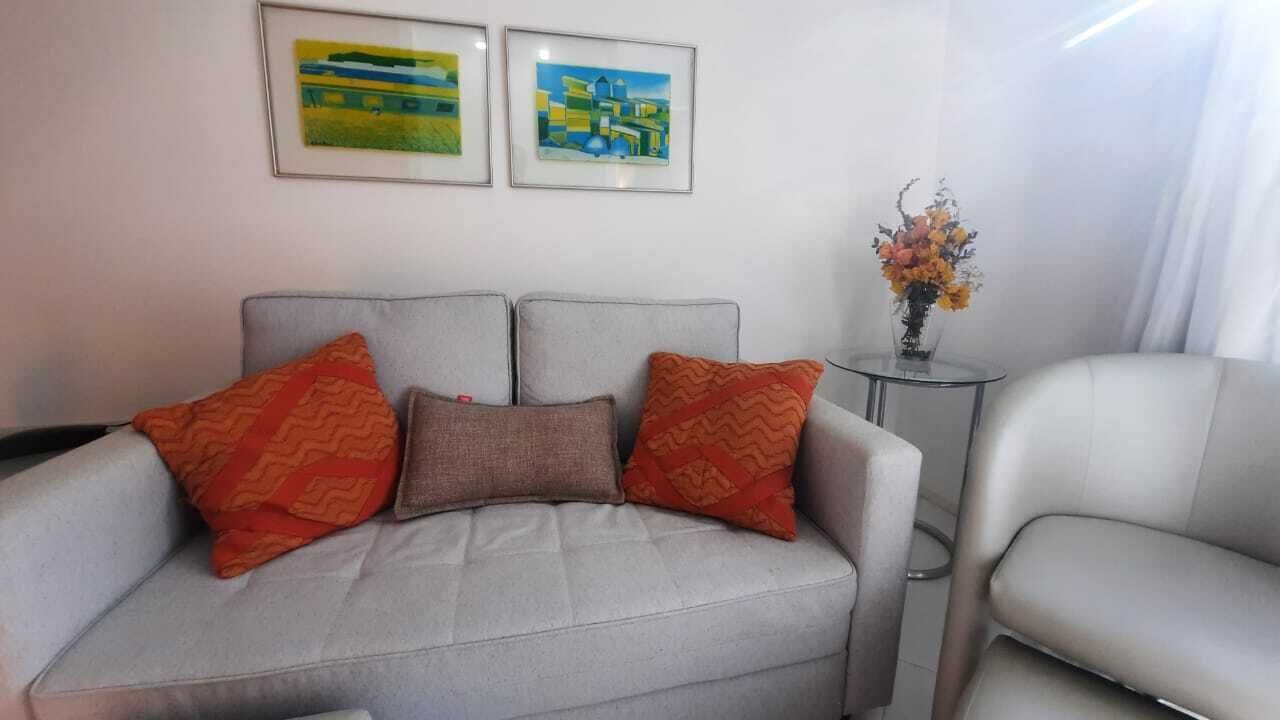 A neutral-colored sofa with patterned cushions, a small side table with a vase of flowers, and two framed artworks on the wall.