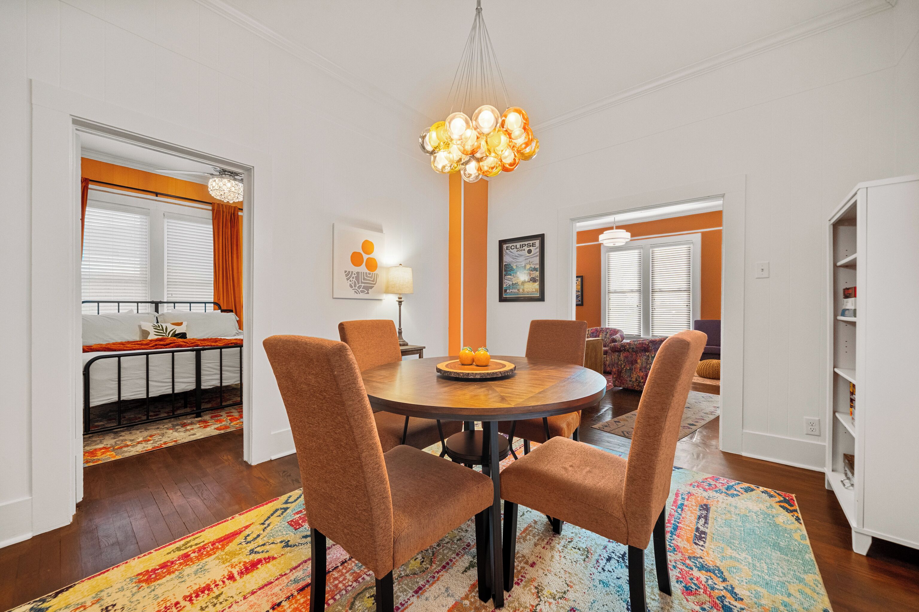 A dining area with a round table, six chairs, a chandelier, and a colorful rug.