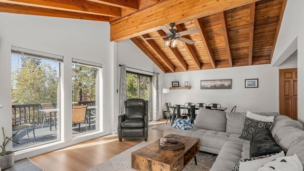 A spacious living room with a high ceiling, a wooden ceiling, a large sofa, a coffee table, and a chair. There is a dining area with a table and chairs, and a large window providing a view of the outdoors. - Tahoe Getaway near Kings Beach - Sleeps 9 (Kings Beach)