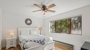 A bedroom with a white bed, a ceiling fan, a wall clock, a bedside table with a lamp, and a window with a view of trees. - Tahoe Getaway near Kings Beach - Sleeps 9 (Kings Beach)