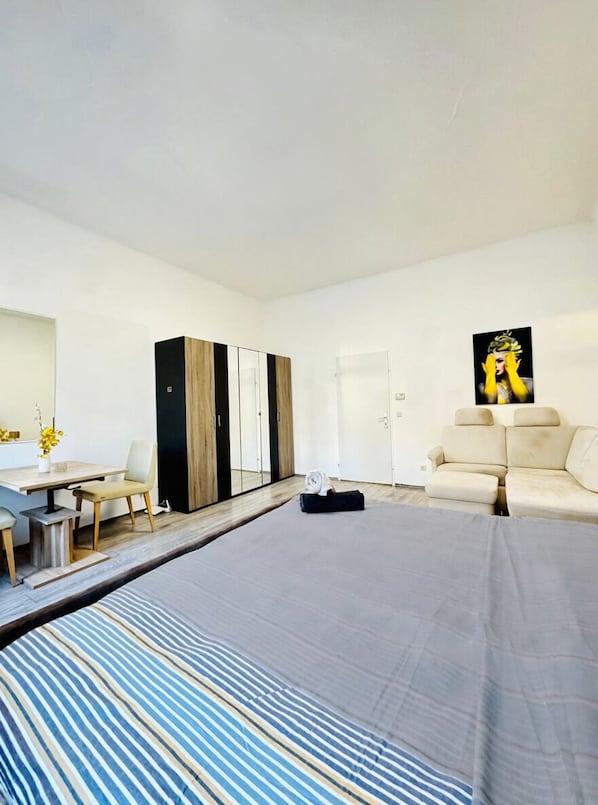 A modern hotel room with a bed, a sofa, a desk with a chair, a wardrobe, and a painting on the wall.