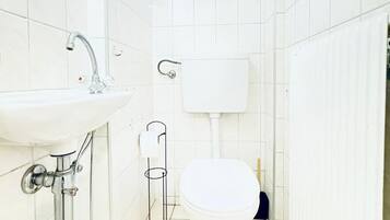Une petite salle de bain blanche, avec des toilettes, un lavabo et une fenêtre agrémentée d’une plante.