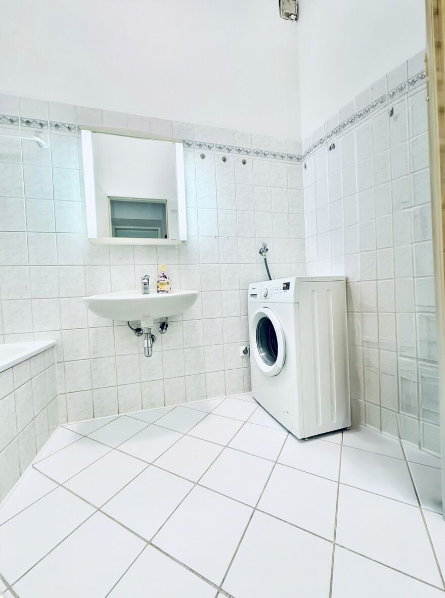 Une petite salle de bain propre, avec des carreaux blancs, un lavabo, un miroir et un lave-linge.