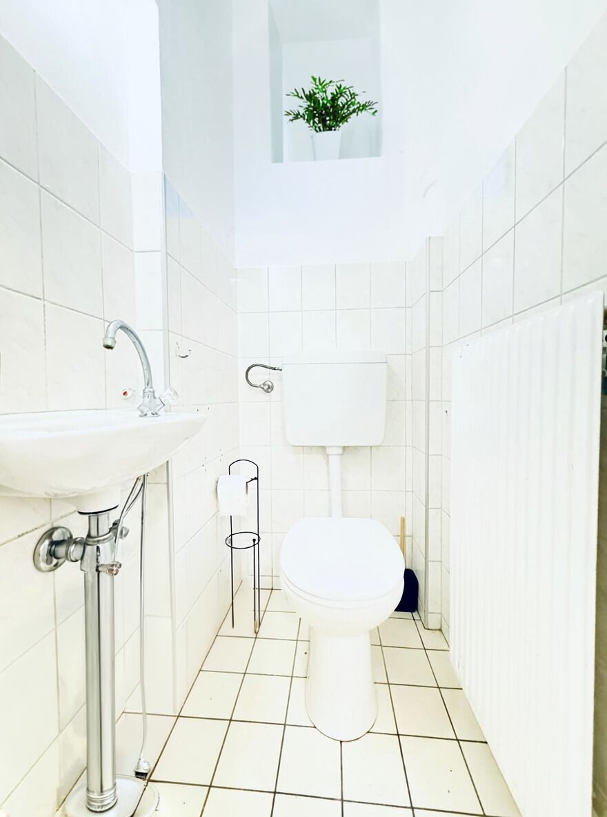 Une petite salle de bain blanche, avec des toilettes, un lavabo et une fenêtre agrémentée d’une plante.