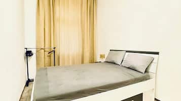 Una cama individual con un colchón gris y dos almohadas, piso de madera, una cortina y una lámpara fijada a la pared.