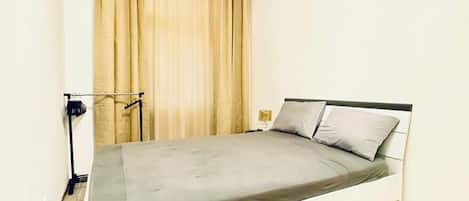 A single bed with a grey mattress and two pillows, a wooden floor, a curtain, and a wall-mounted light fixture.