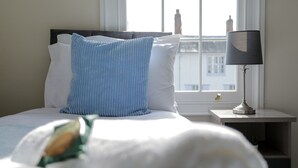 Un lit bien fait, avec un oreiller bleu et des draps blancs, une table de chevet avec une lampe et une fenêtre donnant sur des immeubles.