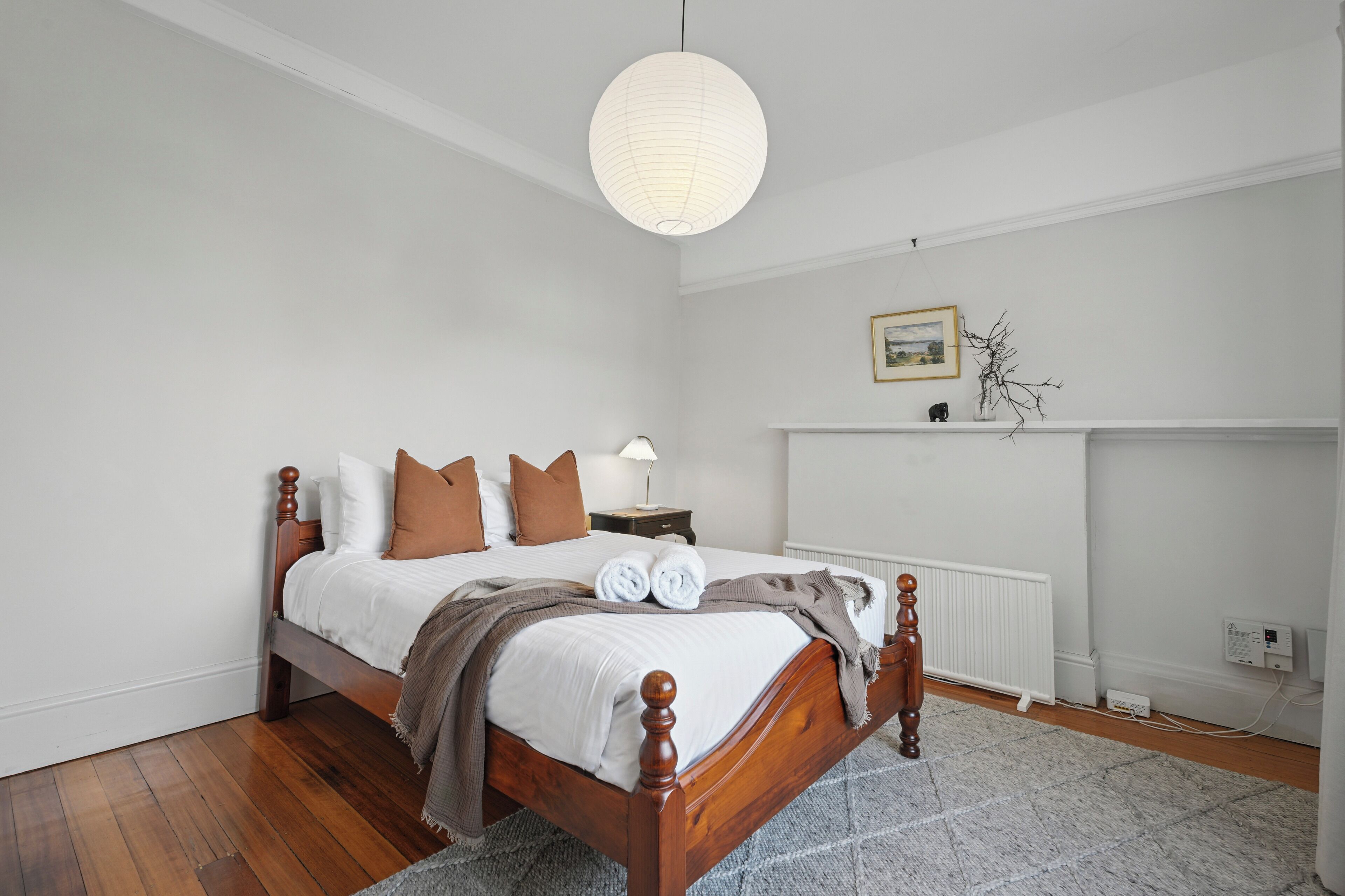 A neatly made bed with a wooden frame, white bedding, and brown pillows. There is a white radiator below the bed, a small bedside table with a lamp, and a picture frame on the wall above the radiator.