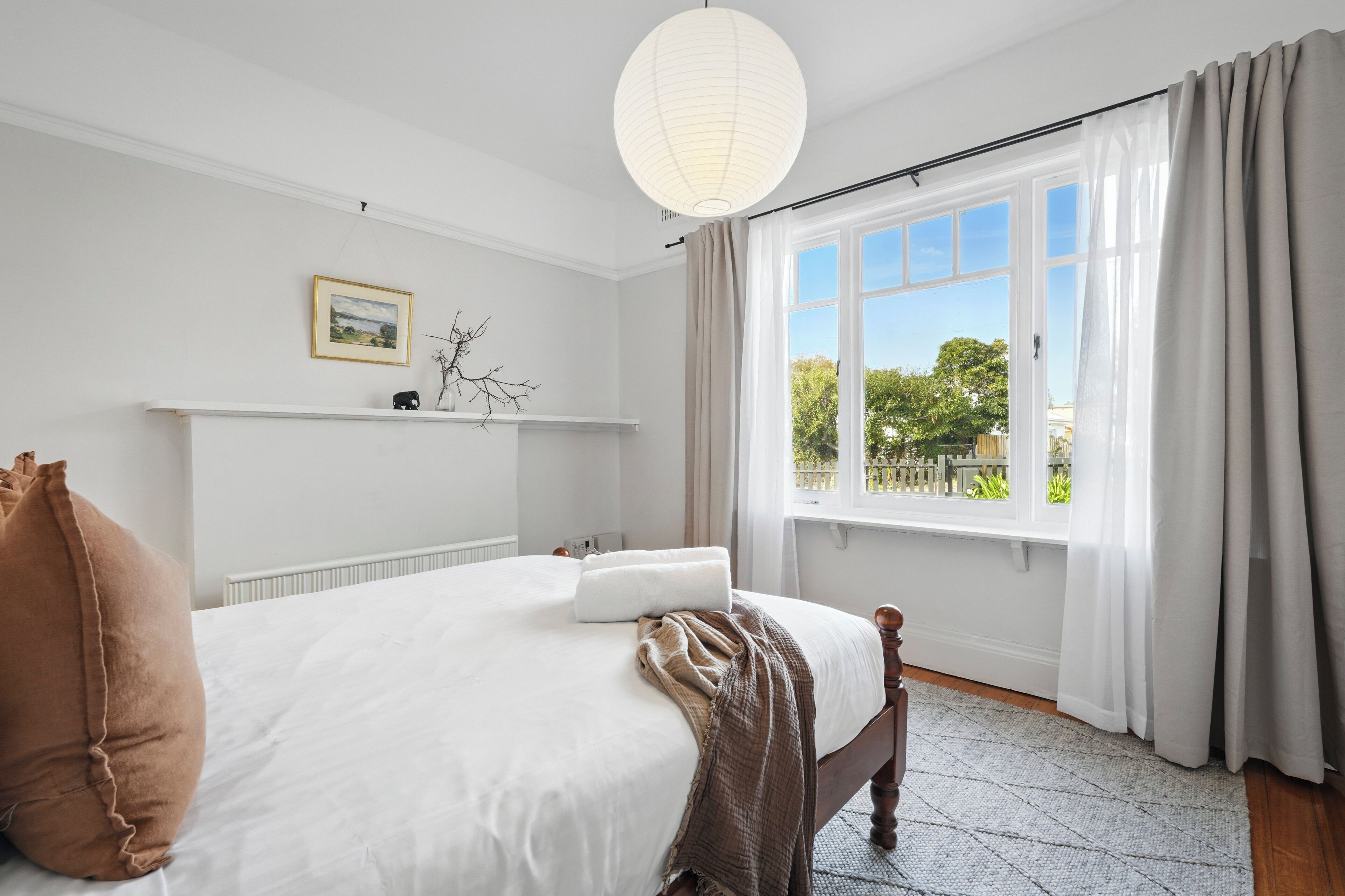 Un dormitorio con una cama, cabecera blanca, ventana con cortinas, radiador y estante en la pared con un cuadro y una planta pequeña.