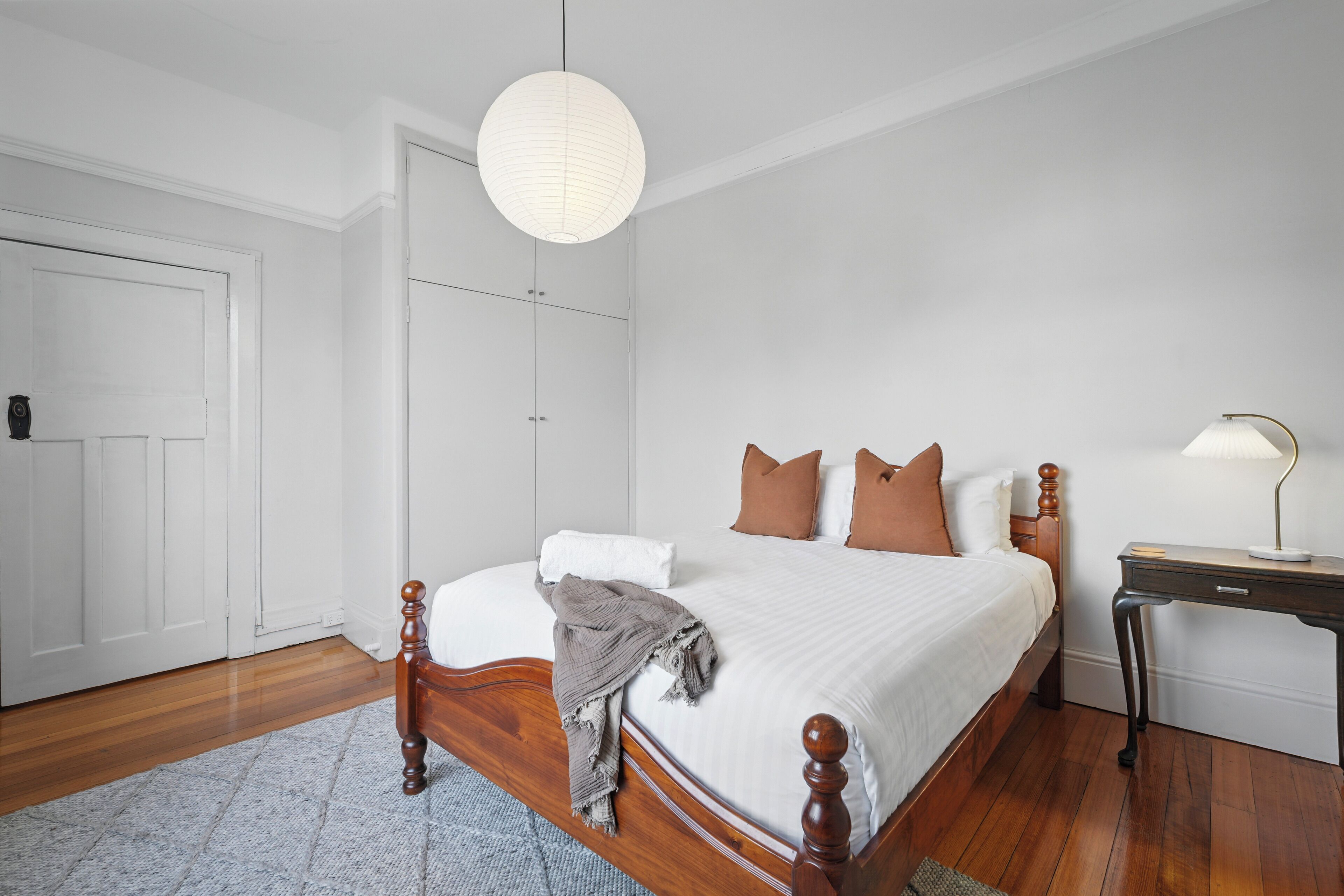 A bedroom with a wooden bed, white walls, a white door, and a bedside table with a lamp.