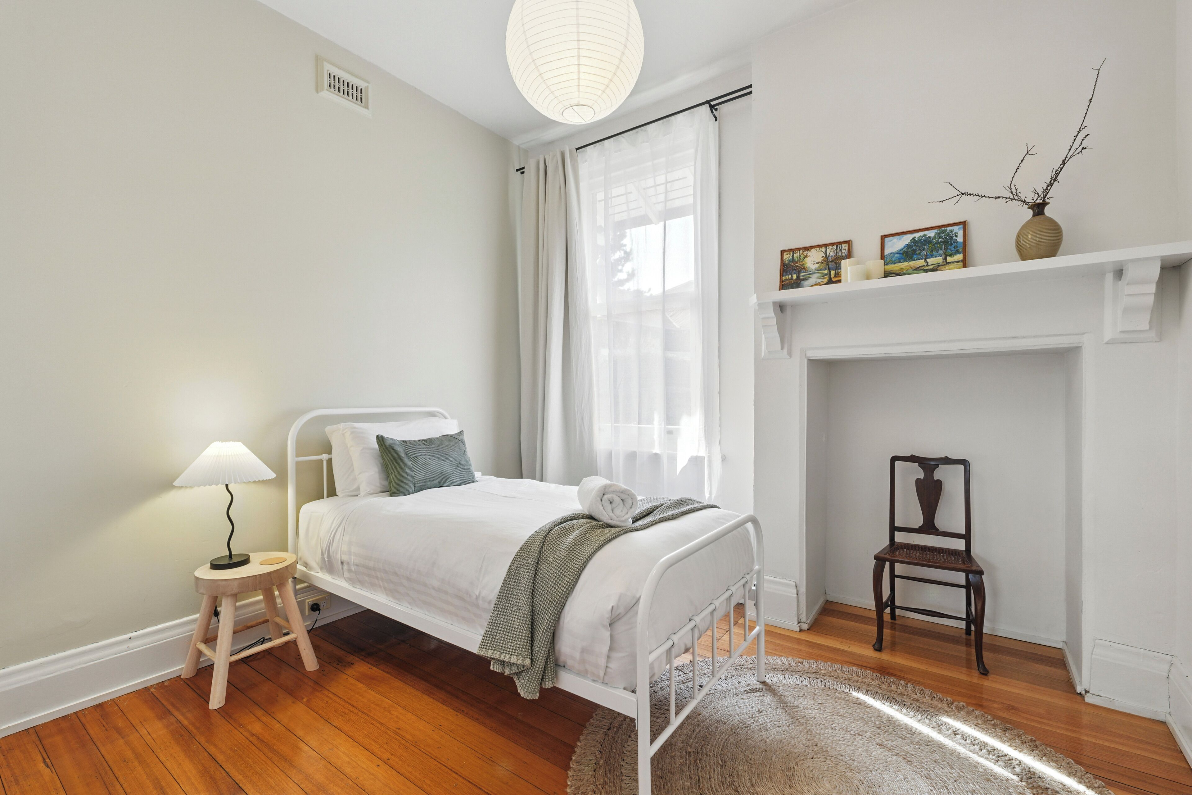 A bedroom with a bed, a small bedside table with a lamp, a chair, a fireplace, and a shelf with pictures and a vase.